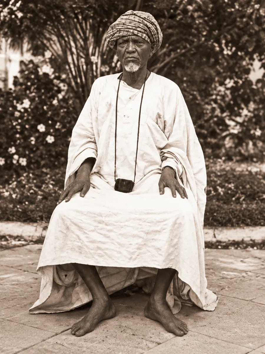 Monsieur Sagou assis dans un jardin, en boubou blanc et coiffe traditionnelle